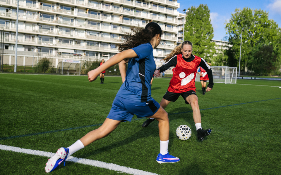 Junger Fußballspieler in blauer Trainingskleidung jongliert einen Fußball auf einem grünen Feld unter klarem Himmel.