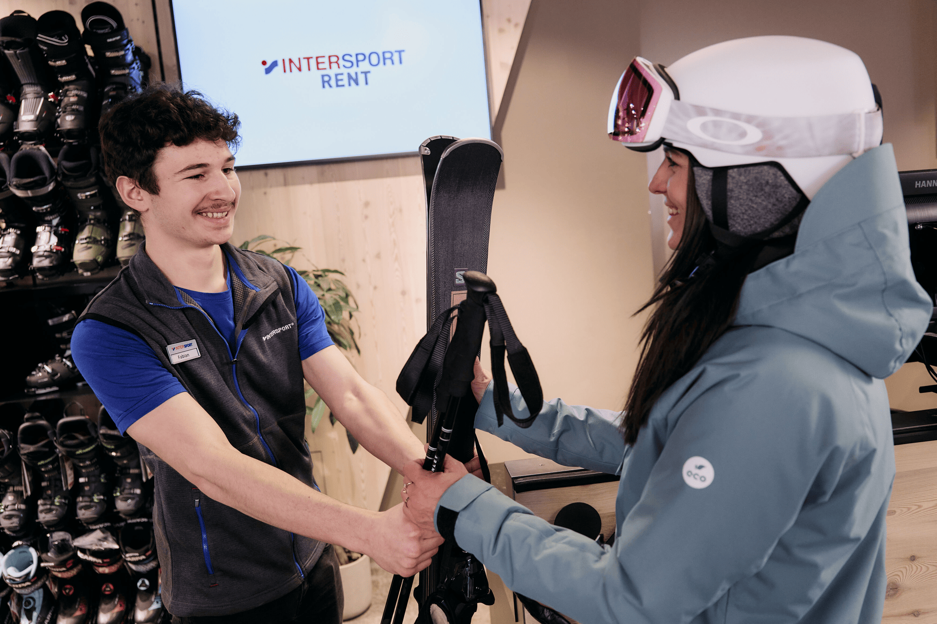 Ein lächelnder Mitarbeiter reicht einer Frau in Skikleidung in einem Verleihgeschäft Skier. An der Wand stehen Skischuhe in Reih und Glied, und ein Schild mit der Aufschrift „Intersport Rent“ ist zu sehen. Die Atmosphäre ist freundlich und hilfsbereit.
