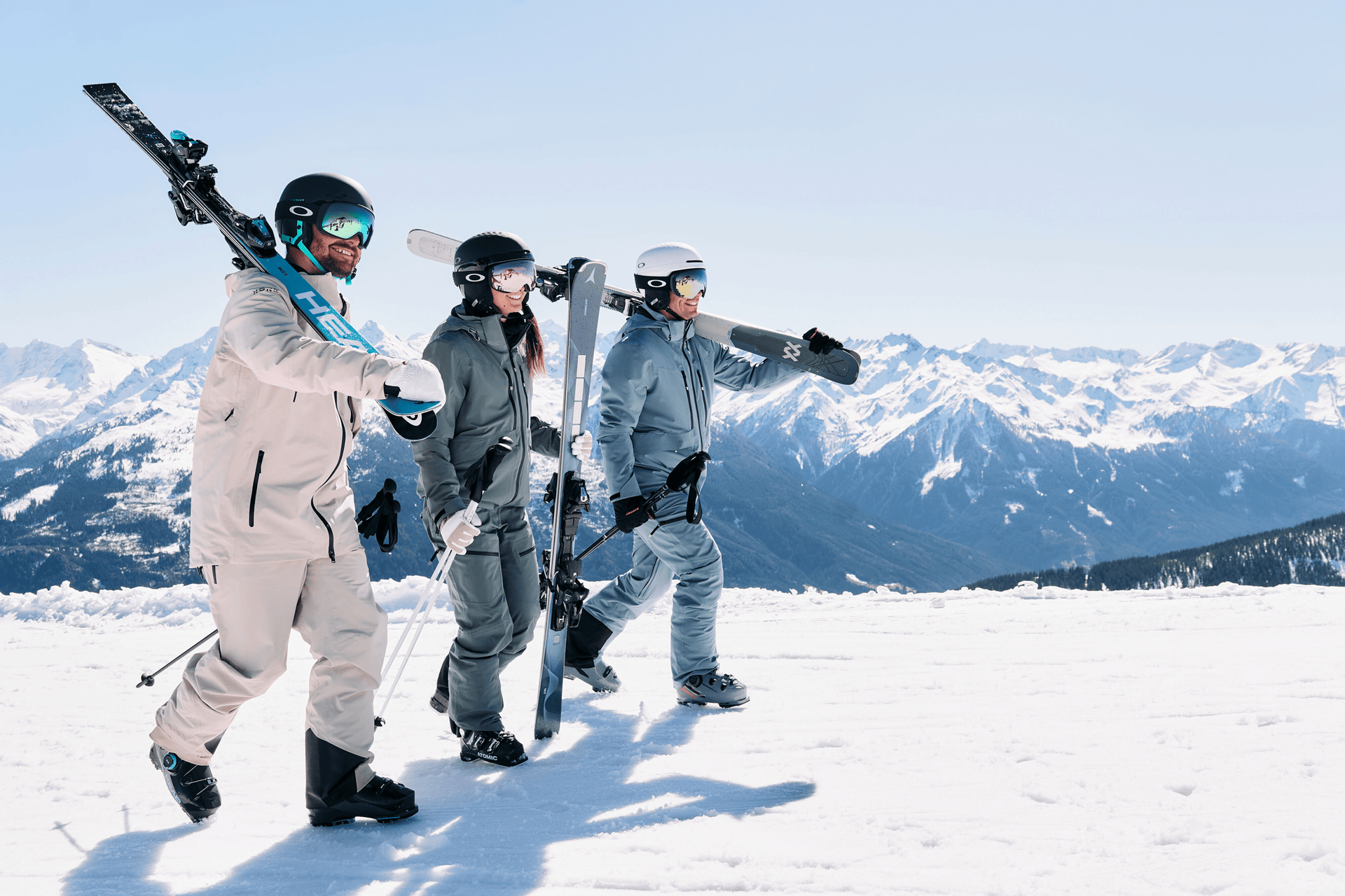 Drei Personen in Skikleidung laufen mit Skiern auf einem schneebedeckten Berg. Der klare blaue Himmel und die majestätischen schneebedeckten Gipfel schaffen eine lebendige, abenteuerliche Atmosphäre.