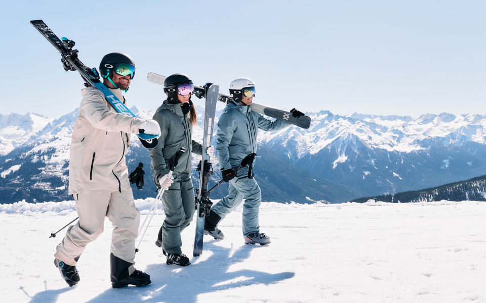 Drei Skifahrer tragen ihre Ski über eine Skipiste und tragen einen Helm, Skianzug und Skischuhe.