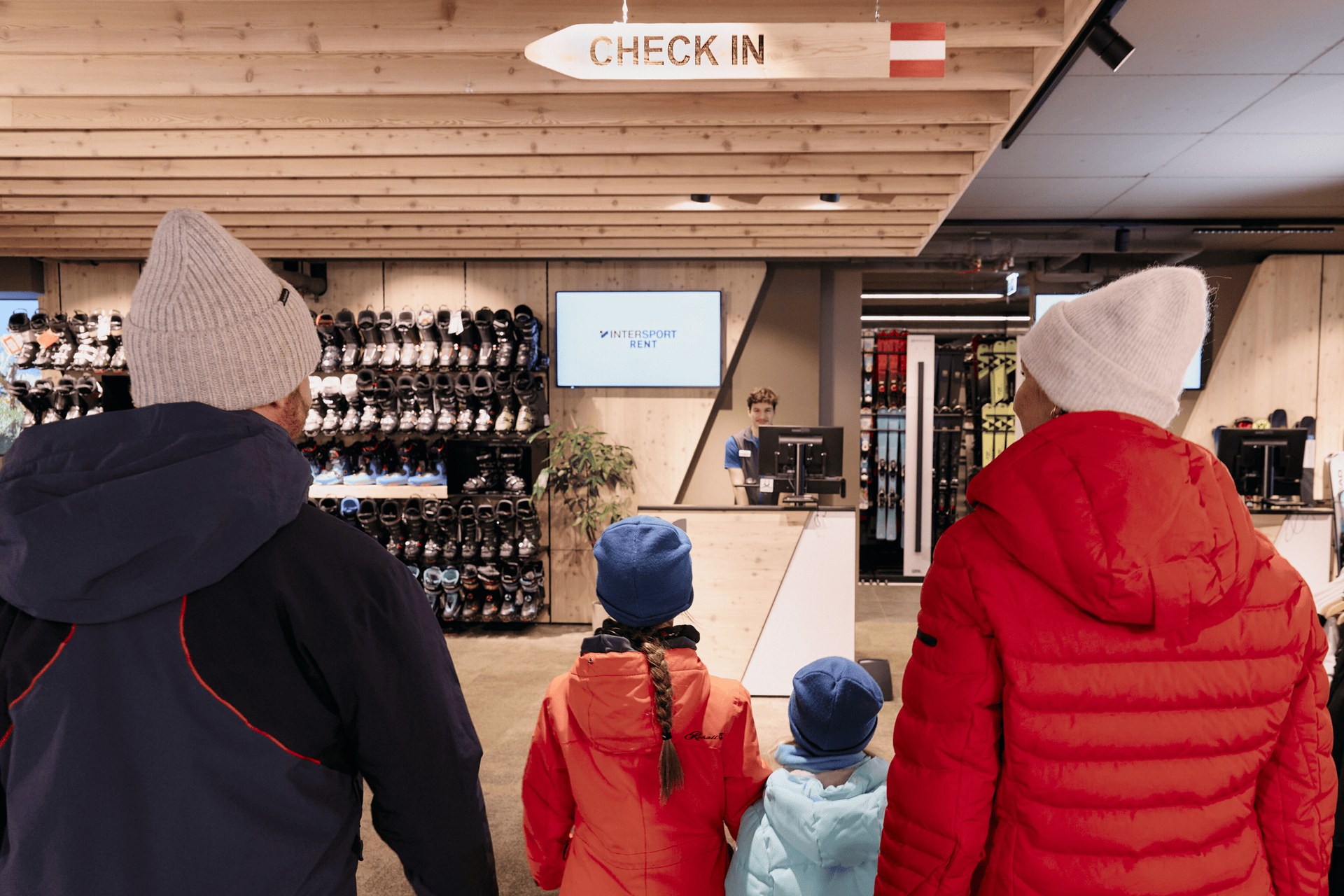 Eine vierköpfige Familie in Winterkleidung steht im Check-in-Bereich eines Skiverleihs. An der Wand reihen sich Regale mit Skischuhen aneinander und schaffen eine gemütliche Atmosphäre.