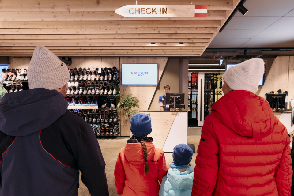 A family of four dressed in winter clothing stands in the check-in area of a ski rental shop. Shelves lined with ski boots line the wall, creating a cozy atmosphere.