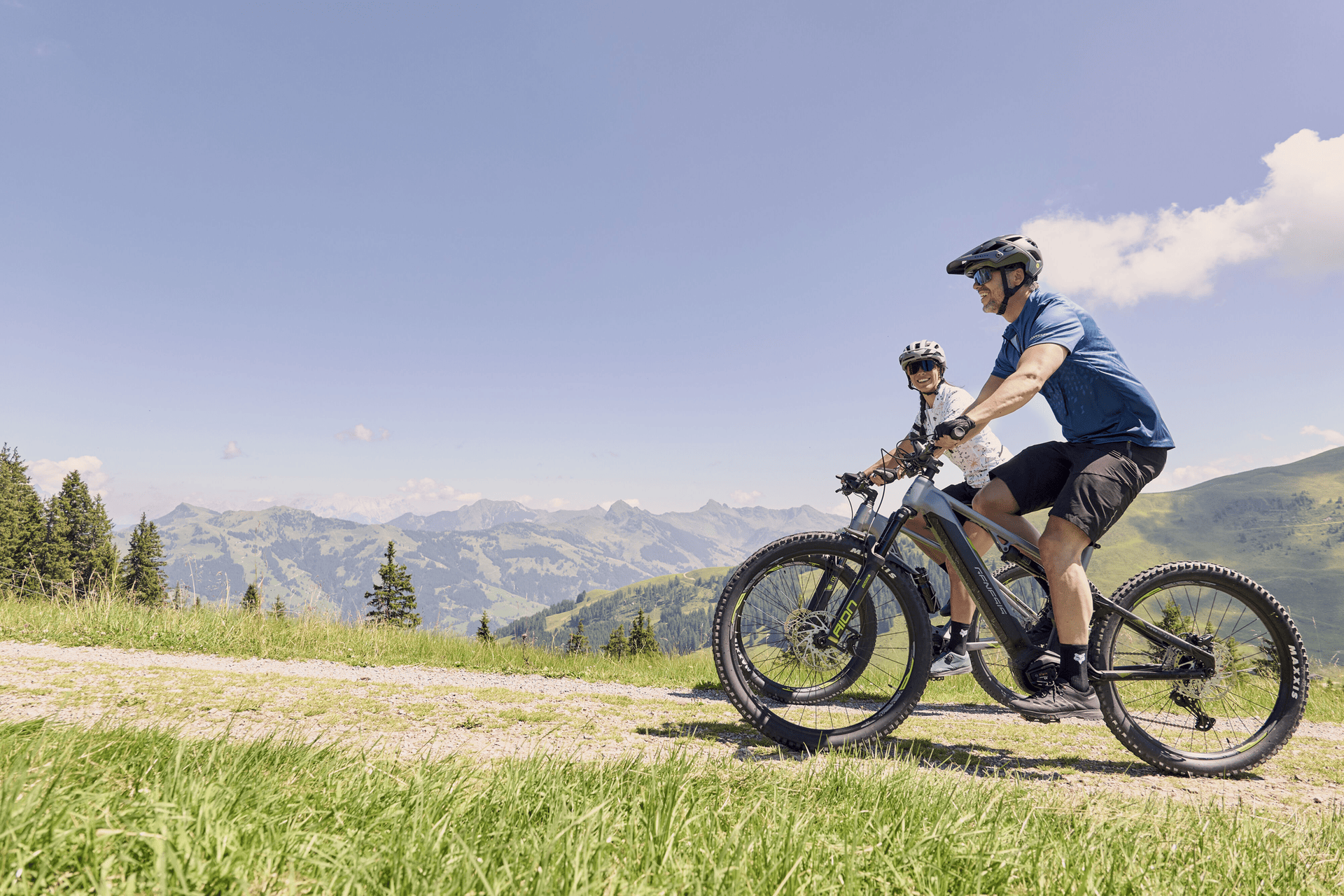Zwei Radfahrer mit Helmen fahren mit E-Bikes auf einem grasbewachsenen Weg mit Bergen im Hintergrund unter einem strahlend blauen Himmel und vermitteln Abenteuerlust und Freude.