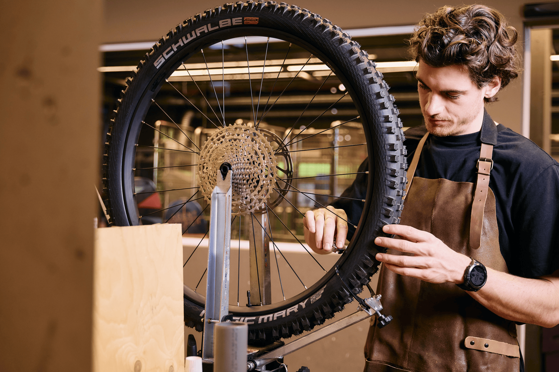 Ein Mann in einer braunen Schürze und einem schwarzen Hemd konzentriert sich in einer Werkstatt auf die Reparatur eines Fahrradrades. Die Umgebung ist gut beleuchtet und detailreich.
