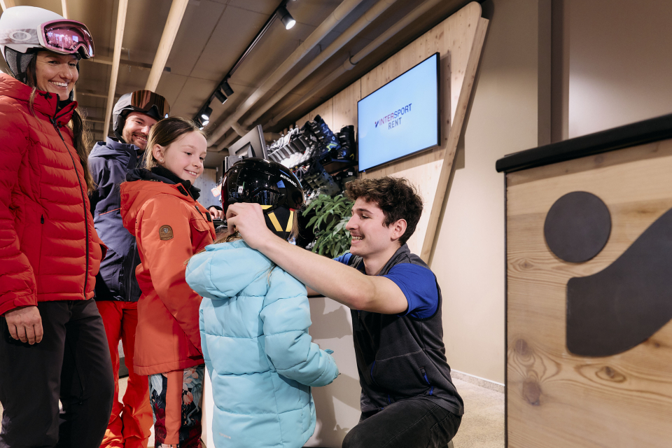 A smiling ski rental staff member helps a young girl with a helmet. Two adults and another child, all in ski gear, watch happily in a cozy shop.