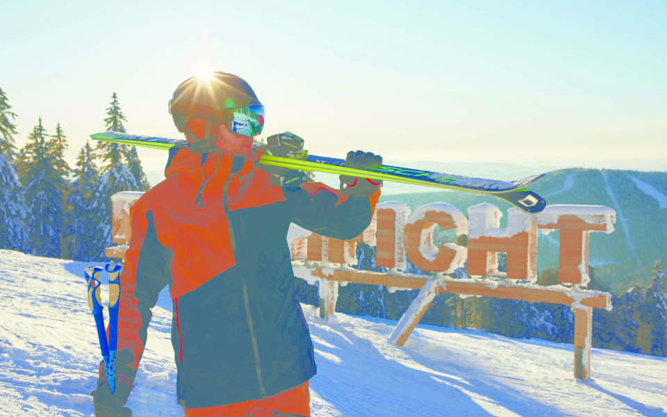 Ein Skifahrer welcher seine Ski auf der Schulter trägt, im Hintergrund befinden sich Holzbuchstaben und schreiben das Wort Hochficht