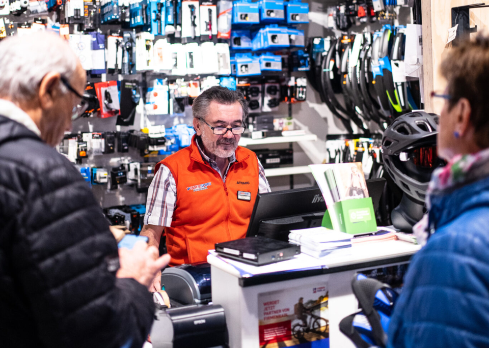 Eine Person kniet nieder und hilft einer anderen Person in roten Hosen und einer grauen Mütze dabei, ihre Skischuhe in einem gemütlichen Skigeschäft mit Holzregalen und Winterausrüstung zu schnüren.