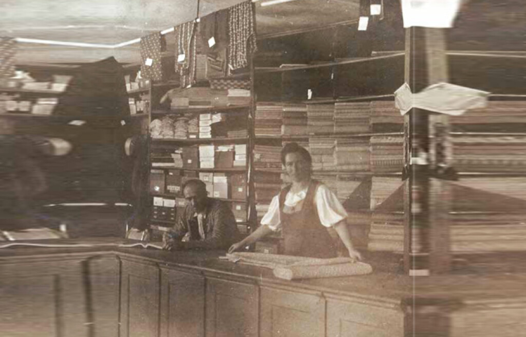 Vintage fotografie obchodu s látkami s prodavači, regály plnými textilií a rolemi látek.