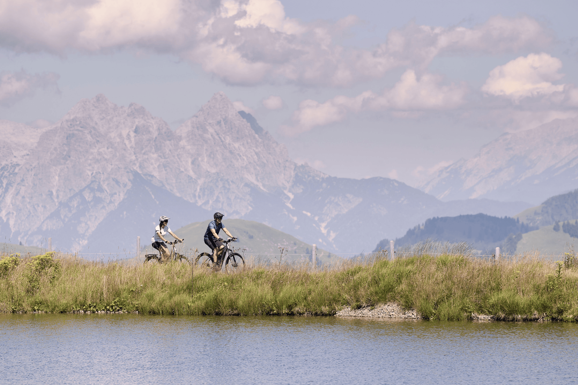 Zwei Radfahrer fahren auf einem grasbewachsenen Weg neben einem ruhigen See entlang, mit majestätischen Bergen im Hintergrund und einem teilweise bewölkten Himmel darüber, was eine friedliche Szenerie schafft.