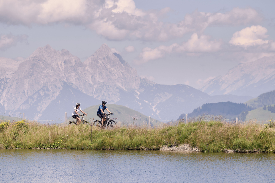 Zwei Radfahrer fahren auf einem grasbewachsenen Weg neben einem ruhigen See entlang, mit majestätischen Bergen im Hintergrund und einem teilweise bewölkten Himmel darüber, was eine friedliche Szenerie schafft.