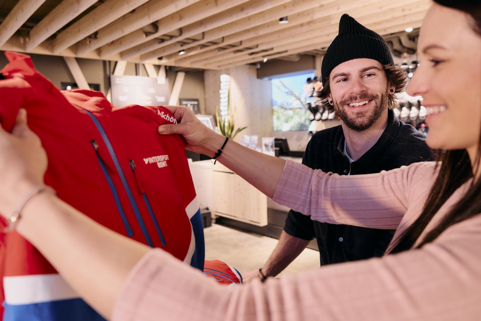 Eine Frau hält in einem Skigeschäft eine rote Jacke mit blauen und weißen Akzenten hoch. Sie lächelt einen Mann mit einer Mütze an. Die Atmosphäre wirkt ungezwungen und freundlich.