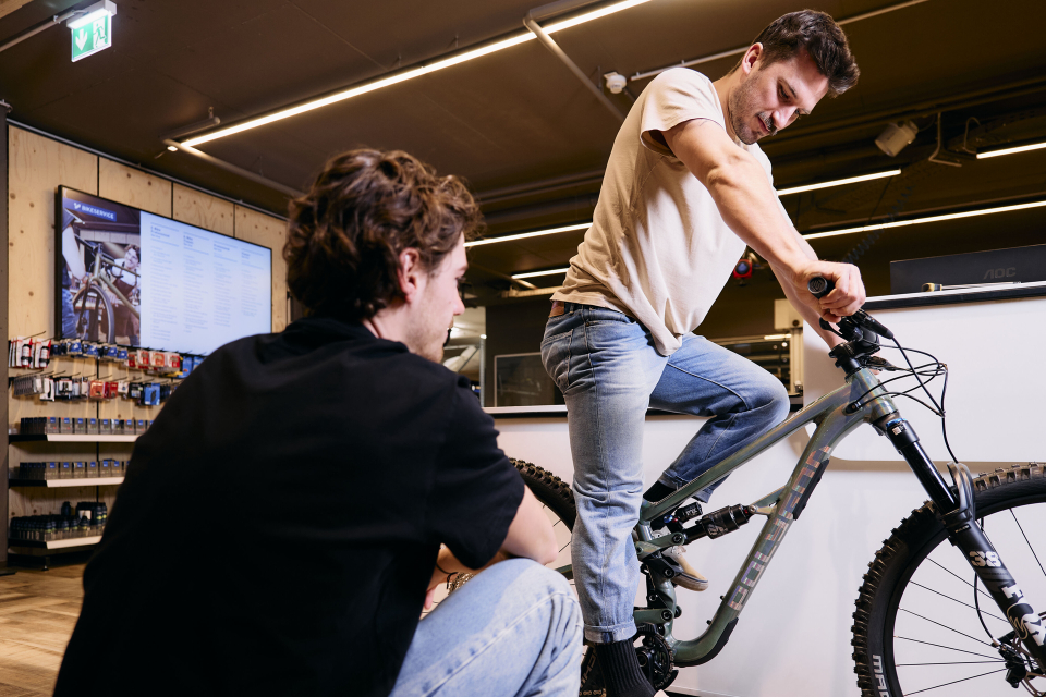 Ein Mann testet ein Mountainbike in einem Innenraum, unterstützt von einer anderen Person, die neben ihm kniet. Der Laden ist modern eingerichtet und präsentiert Fahrradprodukte und einen digitalen Bildschirm.