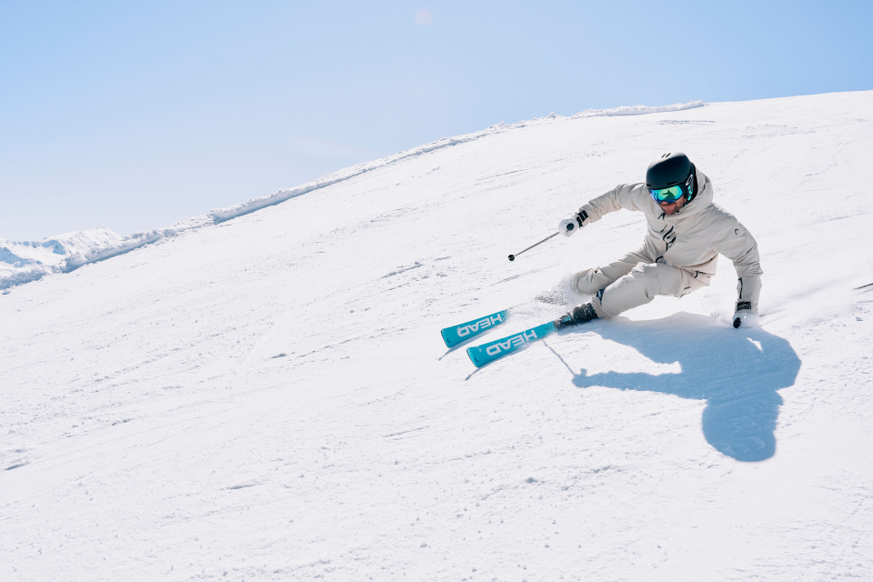 Ein Skifahrer in weißem Anzug und Helm carvt unter strahlend blauem Himmel rasant einen verschneiten Hang hinunter und wirft dabei einen scharfen Schatten auf den unberührten Schnee.