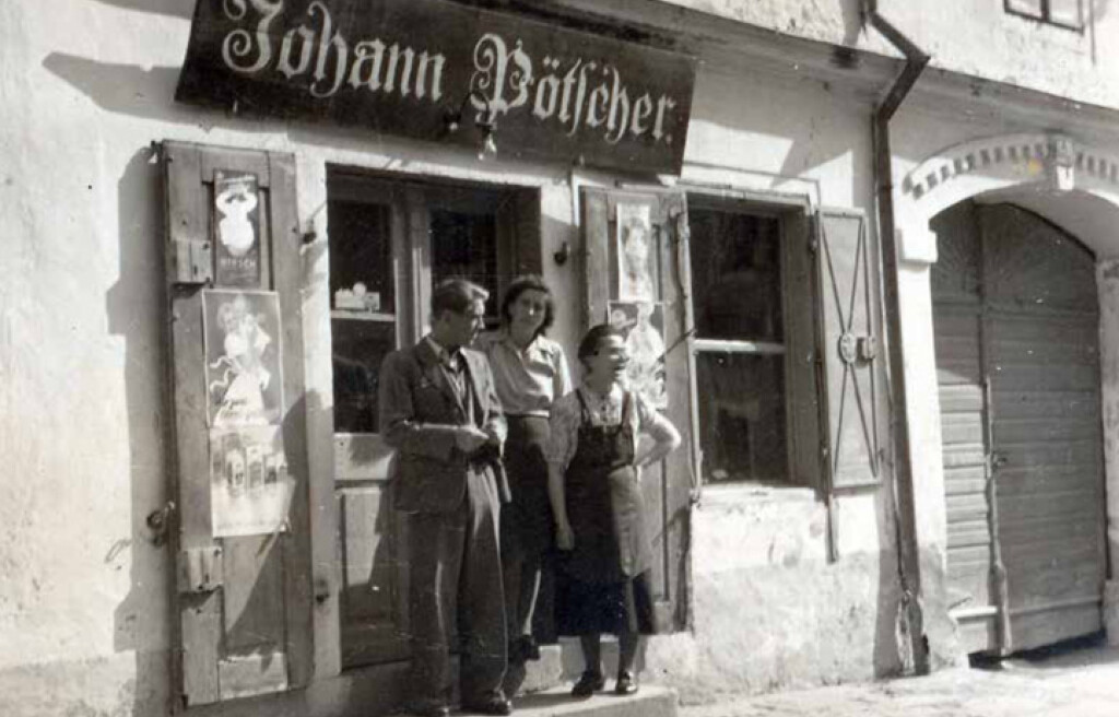 Vintage fotografie exteriéru obchodu Johanna Pötschera se třemi lidmi, kteří stojí venku.