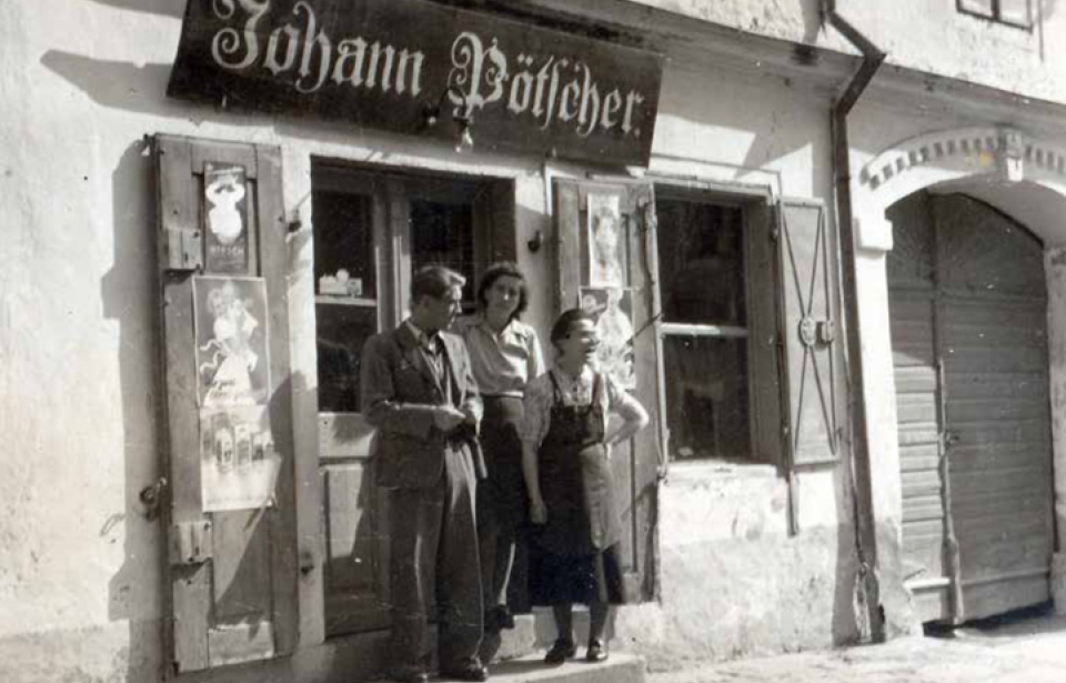 Three people stand in front of a vintage shop with a sign reading "Johann Pofscher." The scene is nostalgic and evokes a sense of mid-20th-century Europe.
