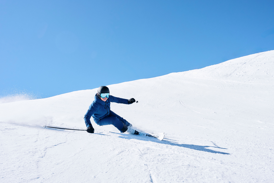 Ein Skifahrer in blauer Kleidung und mit Helm macht eine scharfe Kurve auf einer hellen, verschneiten Piste unter strahlend blauem Himmel und vermittelt dabei Action und Begeisterung.