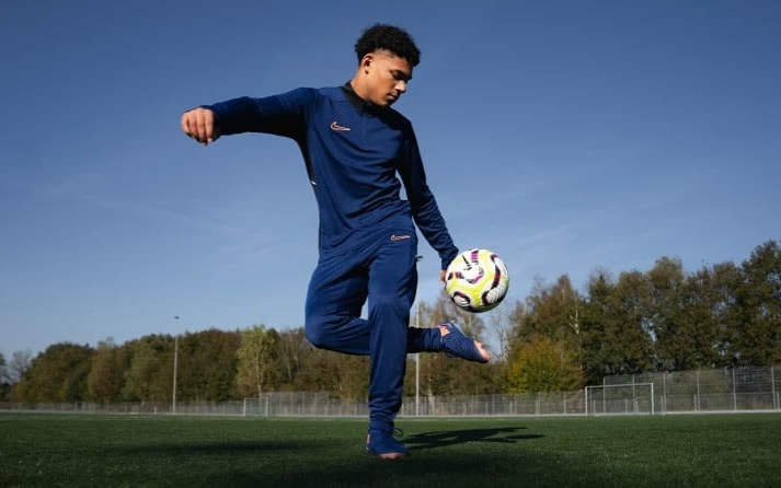 Junger Fußballspieler in blauer Trainingskleidung jongliert einen Fußball auf einem grünen Feld unter klarem Himmel.