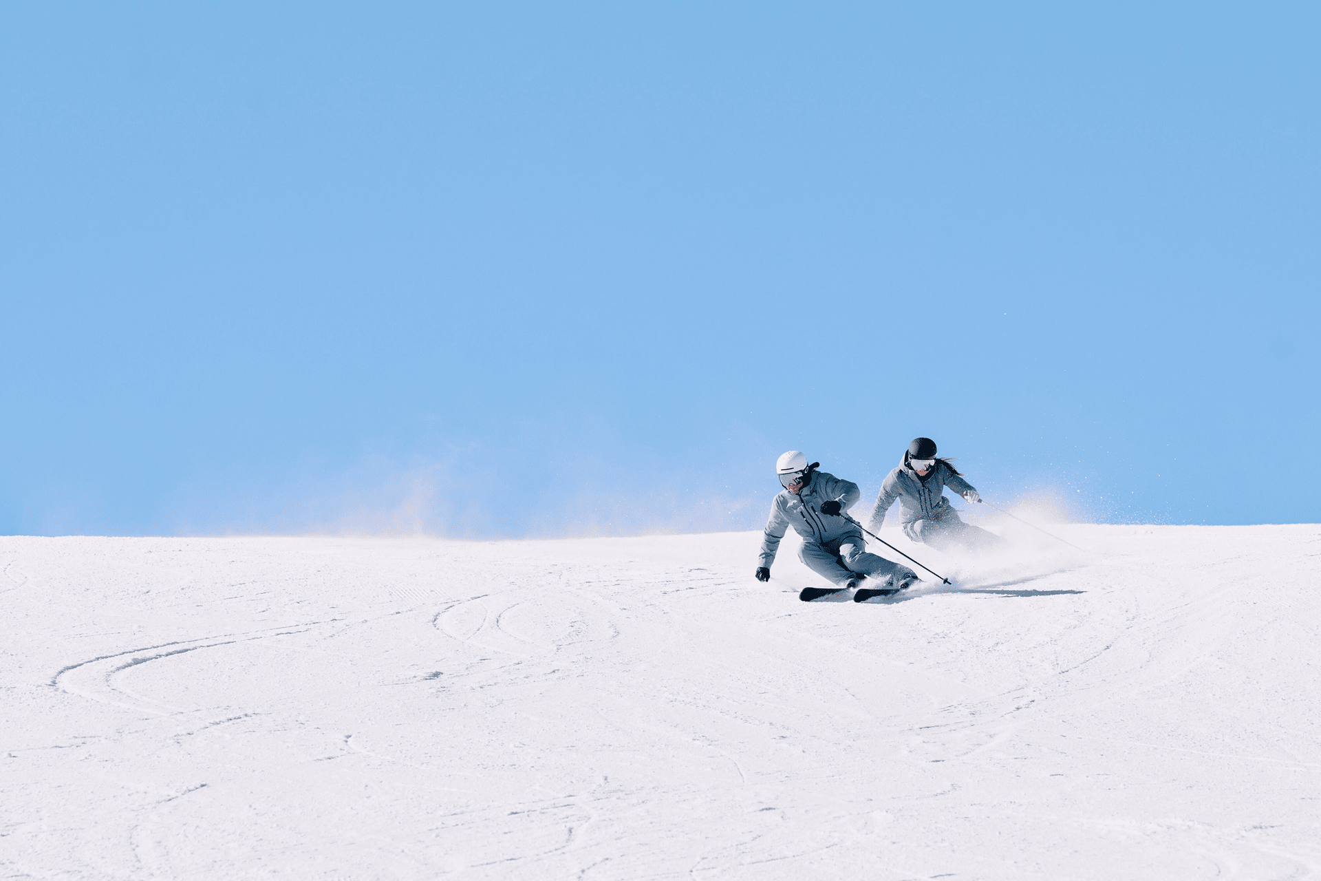 Zwei Skifahrer in Winterausrüstung gleiten vor einem strahlend blauen Himmel schnell einen verschneiten Hang hinunter, wirbeln Pulverschnee auf und strahlen Begeisterung und Energie aus.