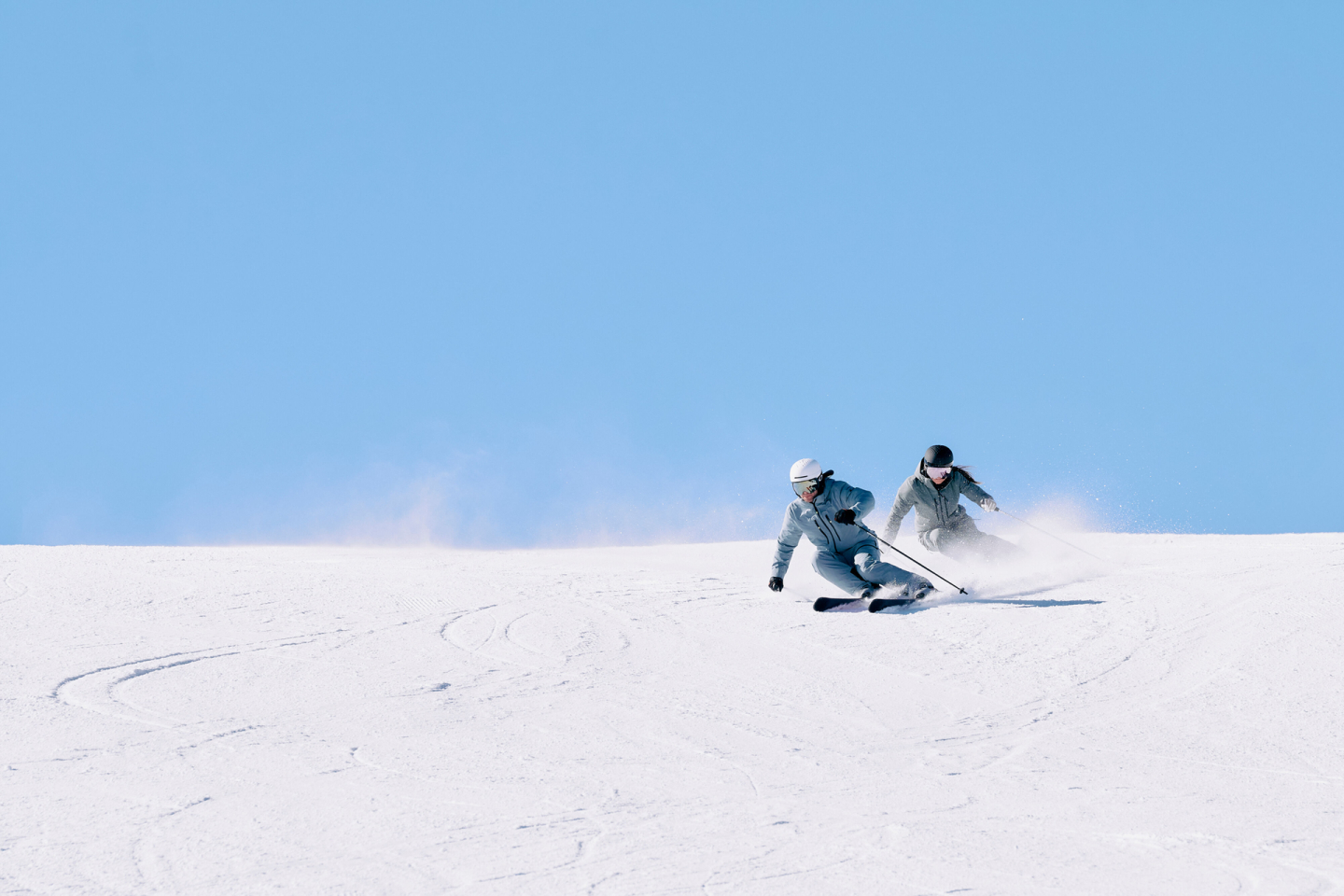 Zwei Skifahrer fahren an einem sonnigen Tag in einem winterlichen Skiresort mit frischem Pulverschnee einen Hang hinunter.
