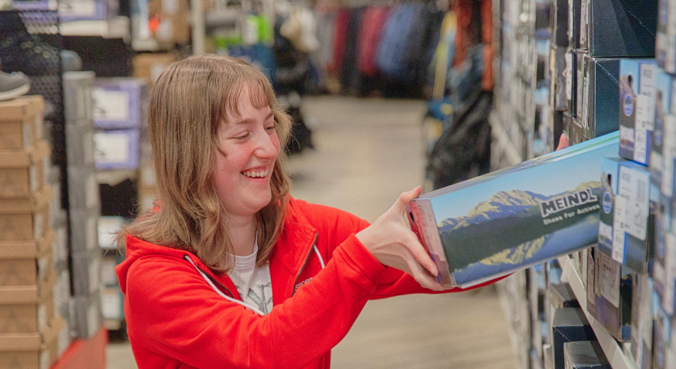 Eine Frau hält in einem Skigeschäft eine rote Jacke mit blauen und weißen Akzenten hoch. Sie lächelt einen Mann mit einer Mütze an. Die Atmosphäre wirkt ungezwungen und freundlich.
