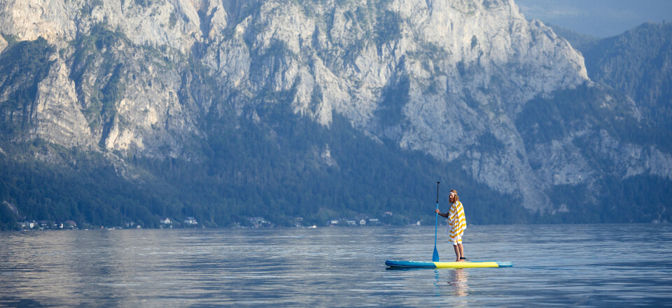 Person stand-up paddleboarding on a calm lake with majestic, tree-covered mountains in the background.