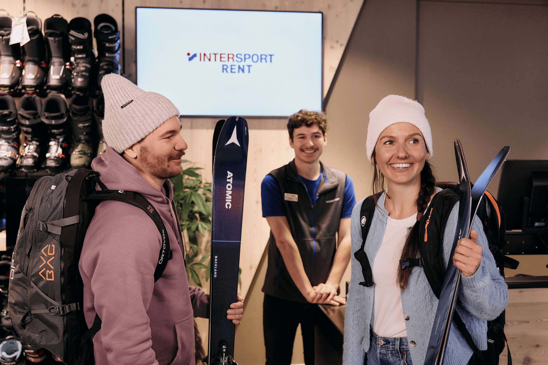 Ein lächelnder Mann und eine lächelnde Frau in Winterkleidung halten Skier in einem Verleihgeschäft in den Händen, hinter ihnen steht ein Mitarbeiter. Skischuhe stehen in den Regalen. Warme, fröhliche Atmosphäre.
