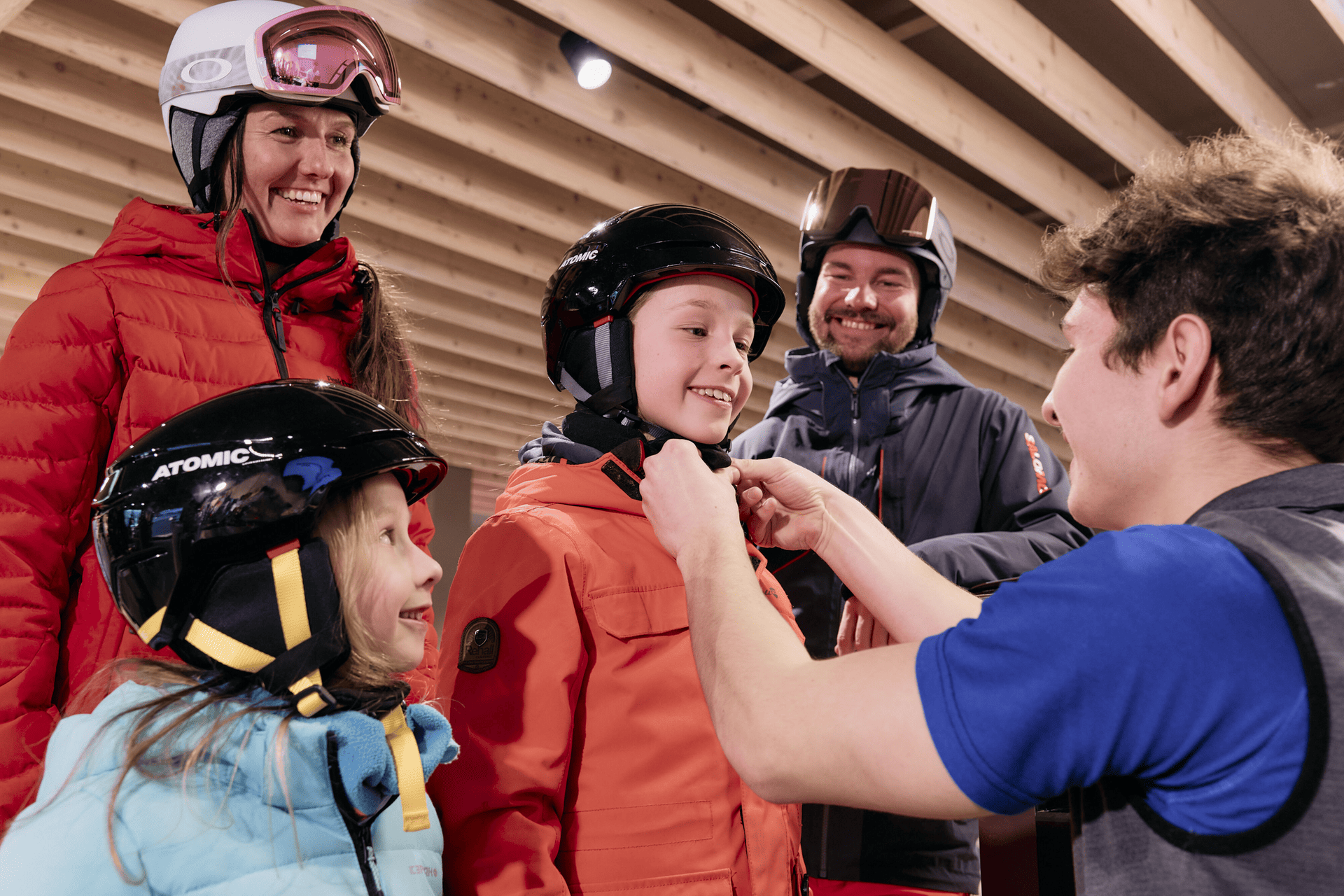 A family is preparing for skiing indoors. The smiling child in orange is fitted with a helmet by a person in blue. The setting is bright and warm.