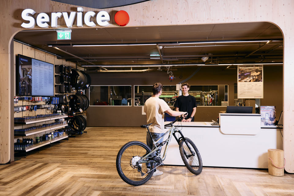 Ein Mann steht mit einem Mountainbike im Servicebereich eines modernen Fahrradgeschäfts. Er scheint sich mit einem Mitarbeiter hinter einem weißen Tresen zu unterhalten. An den Wänden reihen sich Regale und Fahrräder aneinander.