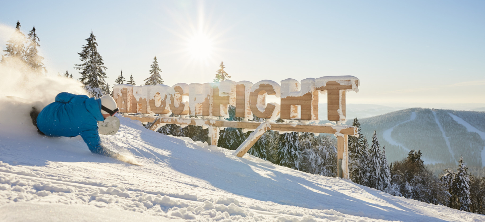 Ein Snowboardfahrer fährt auf einer Skipiste, im Hintergrund befinden sich Holzbuchstaben und schreiben Hochficht