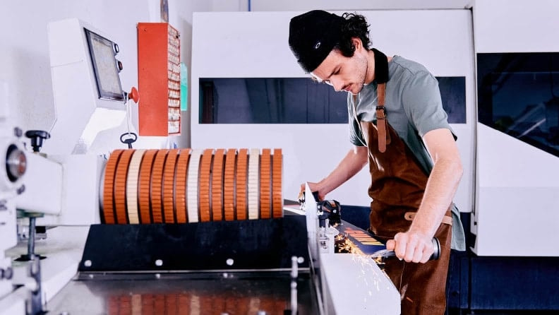 Junger Mann in einer Werkstatt, der die Kante eines Skis an einer Maschine schleift, trägt eine dunkle Mütze und eine braune Schürze, Funken sprühen am Kontaktpunkt.